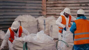 A team of three workers in a white uniform and an orange vest stack and press polyethylene and cellophane while working at a large waste recycling plant video