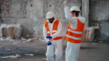 A man with Black skin in a white protective uniform and an orange vest together with his colleague a man with a beard in a white protective helmet dances in the large hall of a waste processing and video