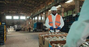 A man with Black skin in a white protective uniform and an orange vest carries boxes with sorted glass garbage and bottles on a special wheelbarrow at a waste processing and sorting plant video