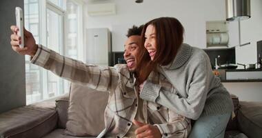 Happy young man with Black skin color in a checkered cream shirt takes a selfie with his brunette girlfriend in a sweater using a white smartphone while sitting on a modern sofa in a studio apartment video