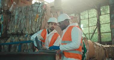 A brunette man with a beard in a white protective suit and an orange vest in a White helmet communicates with a colleague of a man with Black skin who is working on a laptop near huge stacks of video