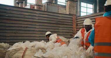 A man with Black skin in a white helmet and a white protective uniform in an orange vest walks on a tramp and presses cellophane garbage along with his colleagues a man with a beard and a girl in a video