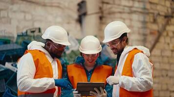 trio of happy successful workers in white uniform and orange vest reviewing their plans on a tablet in a large hall. A man with Black skin a brunette girl and a guy with a beard are talking and video