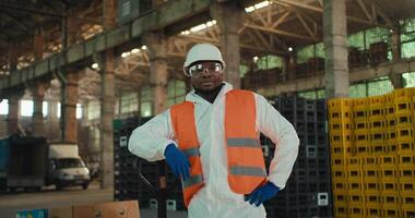 Portrait of a confident man with Black skin in a white protective uniform and an orange vest who stands in safety glasses near large boxes for sorted waste at a plant for processing and sorting video