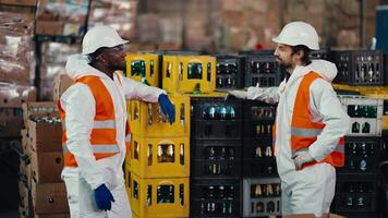 A man with Black skin in a white protective uniform in an orange vest communicates with his male colleague with a beard while they are working and being in a large recycling plant for plastic and video