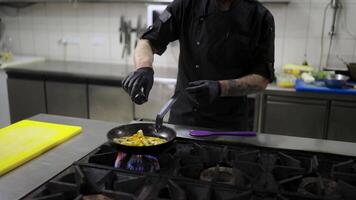 Confident male chef in a black uniform and with black protective gloves pours alcohol onto fried vegetables in a frying pan to apply the flambing technique in a modern kitchen in a restaurant video