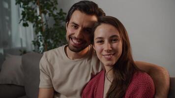 Close-up portrait of a happy young brunette man with stubble hugging his young brunette wife in a red sweater and posing while sitting on a gray sofa in a modern apartment video