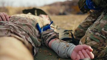 Close-up a military man with his arm bandaged with a cloth bandage lies unconscious on an army stretcher while a combat medic near him looks for a special medicine in his first aid kit during combat video