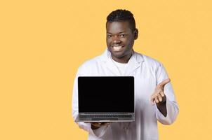 Telemedicine. Toothy black doctor man in white coat uniform showing laptop computer with empty screen, recommending medical website, mockup, standing over yellow studio background photo