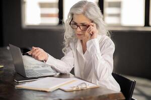 Online Work Routine. Middle Aged Businesswoman Looking At Laptop Through Eyeglasses, Browsing Web And Working Via Computer Sitting In Modern Office. Entrepreneurship Career photo