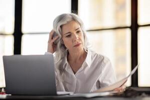 Entrepreneurship Paperwork Problem. Unhappy Businesswoman Looking At Paper Bills And Documents Working In Front Of Laptop Computer, Having Issue With Taxes In Modern Office Indoor photo
