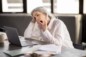 Fatigue Issue. Bored Middle Aged Businesswoman Yawning Looking At Laptop Computer, Tired Of Office Work Routine Sitting At Workplace Indoor. Manager Lady Strugging With Boring Business Tasks photo