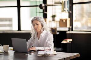 Business Routine. Serious Mature Businesswoman Typing On Laptop Computer, Solving Issues Sitting In Modern Office. Manager Lady Focused On Difficult Task Browsing Internet At Workplace Indoor photo