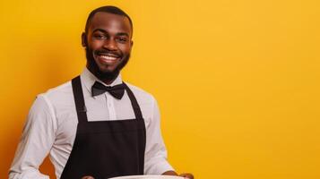 A cheerful waiter wearing a black apron and bow tie holds a plate, standing in front of a vibrant yellow background. photo