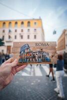 Hand holding Colosseum postcard in Rome, Italy. Seamless transition from postcard to real buildings creating intriguing visual effect. Urban setting with European architectural style. photo