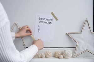 Unrecognizable woman creating vision board. NEW YEAR'S RESOLUTION checklist text on paper note stick on while wall with cozy minimalistic handmade Christmas decor. New year aims resolutions. Low key festive Planning and setting goals concept photo