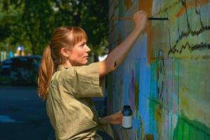 Pretty graffiti artist painter with a brush on a stepladder paints a wall photo