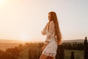 Happy woman in white boho dress on sunset in mountains. Romantic woman with long hair standing with her back on the sunset in nature in summer with open hands. Silhouette. Nature. Sunset. photo