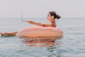Woman freelancer works on laptop swimming in sea on pink inflatable ring. Happy tourist floating on inflatable donut and working on laptop computer in calm ocean. Freelance, remote working anywhere photo