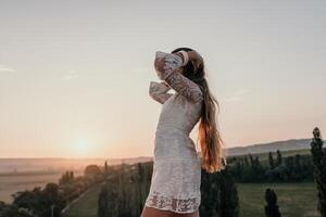 Happy woman in white boho dress on sunset in mountains. Romantic woman with long hair standing with her back on the sunset in nature in summer with open hands. Silhouette. Nature. Sunset. photo