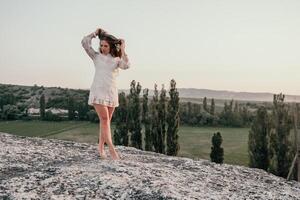 Happy woman in white boho dress on sunset in mountains. Romantic woman with long hair standing with her back on the sunset in nature in summer with open hands. Silhouette. Nature. Sunset. photo