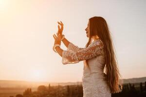Happy woman in white boho dress on sunset in mountains. Romantic woman with long hair standing with her back on the sunset in nature in summer with open hands. Silhouette. Nature. Sunset. photo