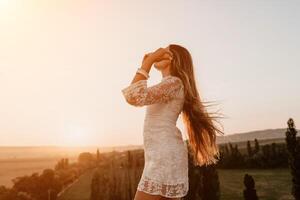 Happy woman in white boho dress on sunset in mountains. Romantic woman with long hair standing with her back on the sunset in nature in summer with open hands. Silhouette. Nature. Sunset. photo
