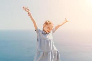 A young girl is standing on a beach, wearing a blue dress and holding her arms up in the air. Concept of joy and freedom, as the girl appears to be enjoying her time by the ocean. photo