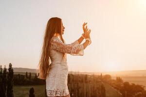 Happy woman in white boho dress on sunset in mountains. Romantic woman with long hair standing with her back on the sunset in nature in summer with open hands. Silhouette. Nature. Sunset. photo