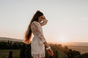 Happy woman in white boho dress on sunset in mountains. Romantic woman with long hair standing with her back on the sunset in nature in summer with open hands. Silhouette. Nature. Sunset. photo