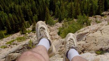 Man is looking down at his feet as he sits on the edge of the cliff, surrounded by mountains, rivers, and forests at the summit. video