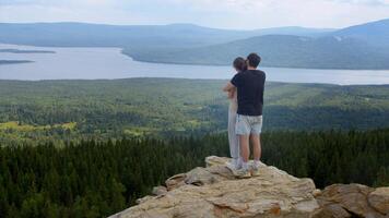 Young man stands behind his girlfriend on a mountain top next to a cliff. He gives her a hug and shows her the beautiful view of the mountains, forests, and winding river. video