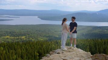 Two lovers, holding hands and kissing, stand by a cliff at the top of a mountain, with a beautiful natural landscape as a backdrop. video