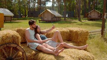 Couple takes a selfie sitting on some hay, with different summer houses as a backdrop, in a camping setting. video