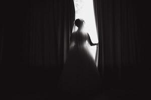 Portrait of beautiful bride in white silk dressing gown with curly hairstyle and veil standing near window in bedroom, copy space. Bride's morning preparation. Back view photo