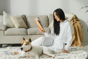 Young smiling woman resting on sofa and reading a book with dog sitting near by her at home. photo