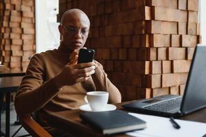 African businessman talking on phone sitting at cafe table, busy entrepreneur working distantly in coffee house with laptop papers speaking on mobile, black man making call having lunch in cafeteria. photo