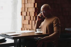 African businessman talking on phone sitting at cafe table, busy entrepreneur working distantly in coffee house with laptop papers speaking on mobile, black man making call having lunch in cafeteria. photo