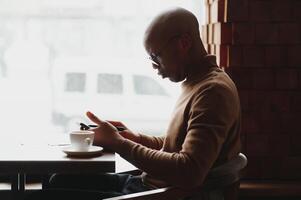 African businessman talking on phone sitting at cafe table, busy entrepreneur working distantly in coffee house with laptop papers speaking on mobile, black man making call having lunch in cafeteria. photo