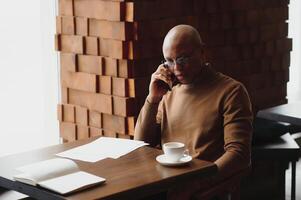 African businessman talking on phone sitting at cafe table, busy entrepreneur working distantly in coffee house with laptop papers speaking on mobile, black man making call having lunch in cafeteria. photo