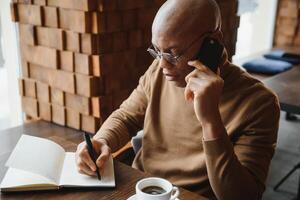African businessman talking on phone sitting at cafe table, busy entrepreneur working distantly in coffee house with laptop papers speaking on mobile, black man making call having lunch in cafeteria. photo
