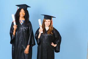 Two girls are posing for take photo in black gowns and hold diploma certificate. They are graduates and hold diploma certificate. They are happy and in good mood.