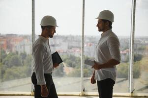A front view of two smart architects with white helmets reviewing blueprints at a construction site on a bright sunny day photo