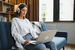 Smiling attractive young woman sitting on sofa using laptop communicating working online at home, happy teen girl typing on computer, enjoying writing blog or chatting with friends in social network photo