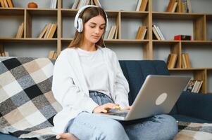 Smiling attractive young woman sitting on sofa using laptop communicating working online at home, happy teen girl typing on computer, enjoying writing blog or chatting with friends in social network photo