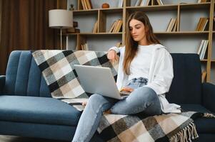 Smiling attractive young woman sitting on sofa using laptop communicating working online at home, happy teen girl typing on computer, enjoying writing blog or chatting with friends in social network photo