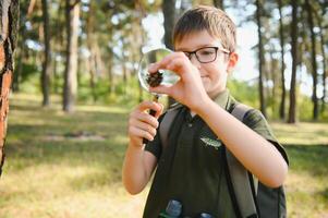 schoolboy is exploring nature with magnifying glass. Summer vacation for inquisitive kids in forest. Hiking. Boy-scout. photo