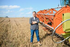 Happy farmer proudly standing in a field. Combine harvester driver going to crop rich wheat harvest. Agronomist wearing flannel shirt, looking at camera on a farmland photo