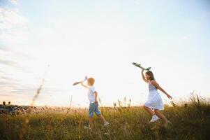 Running boy and girl holding two green and blue airplanes toy in the field during summer sunny day photo