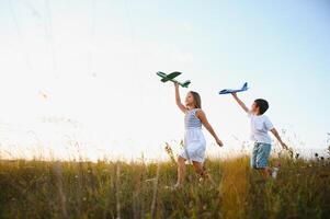 Running boy and girl holding two green and blue airplanes toy in the field during summer sunny day photo